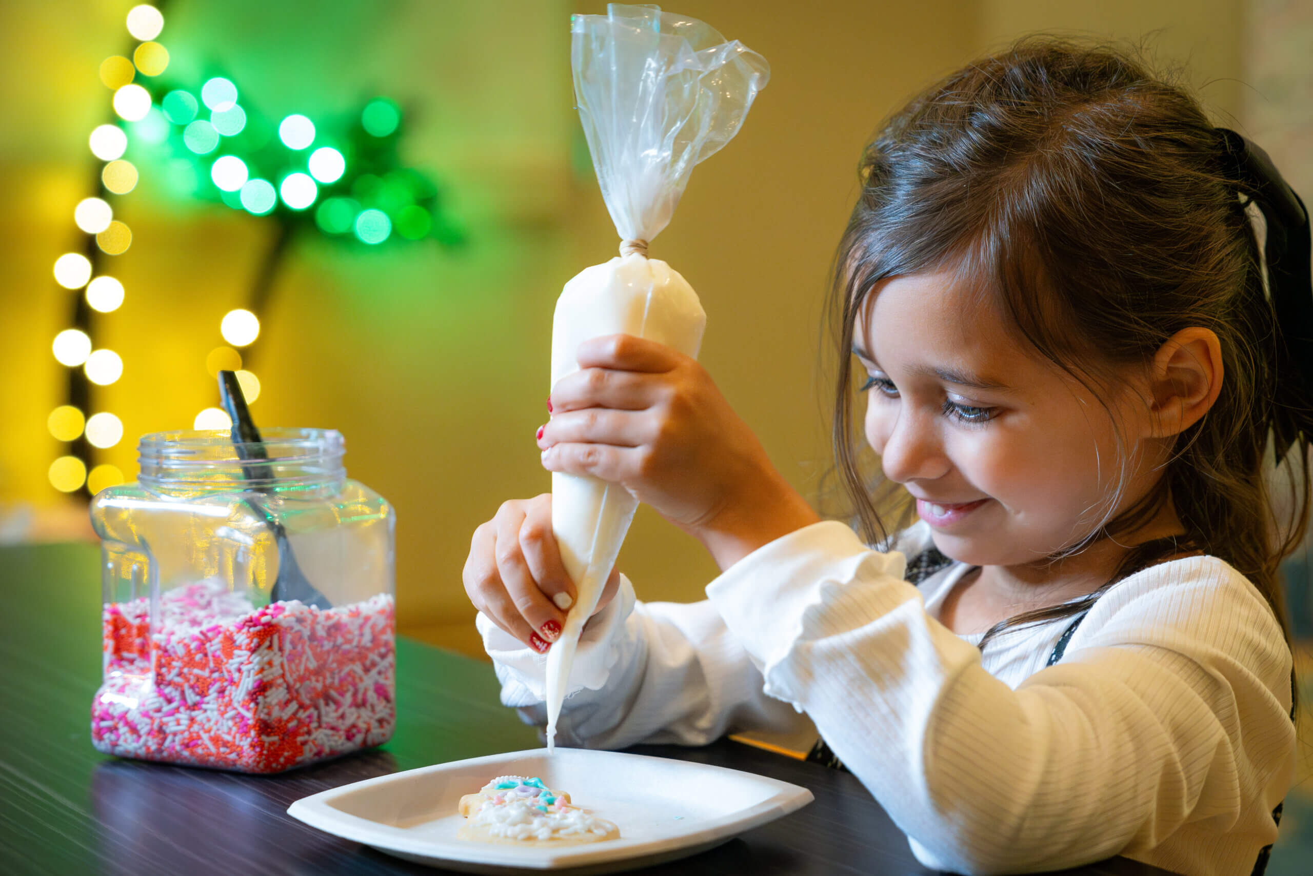 A young girl smiles while decorating a cookie with white icing from a piping bag. A jar of colorful sprinkles sits nearby on the table, and festive lights glow in the blurred background.