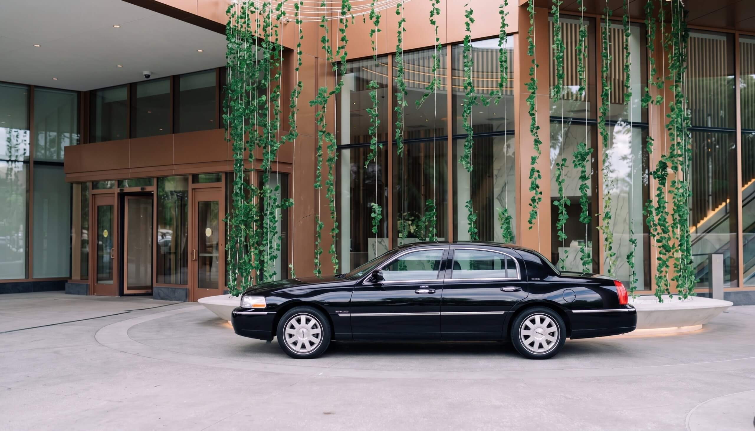 A black sedan is parked in front of a modern building with large windows and hanging green vines as decoration.