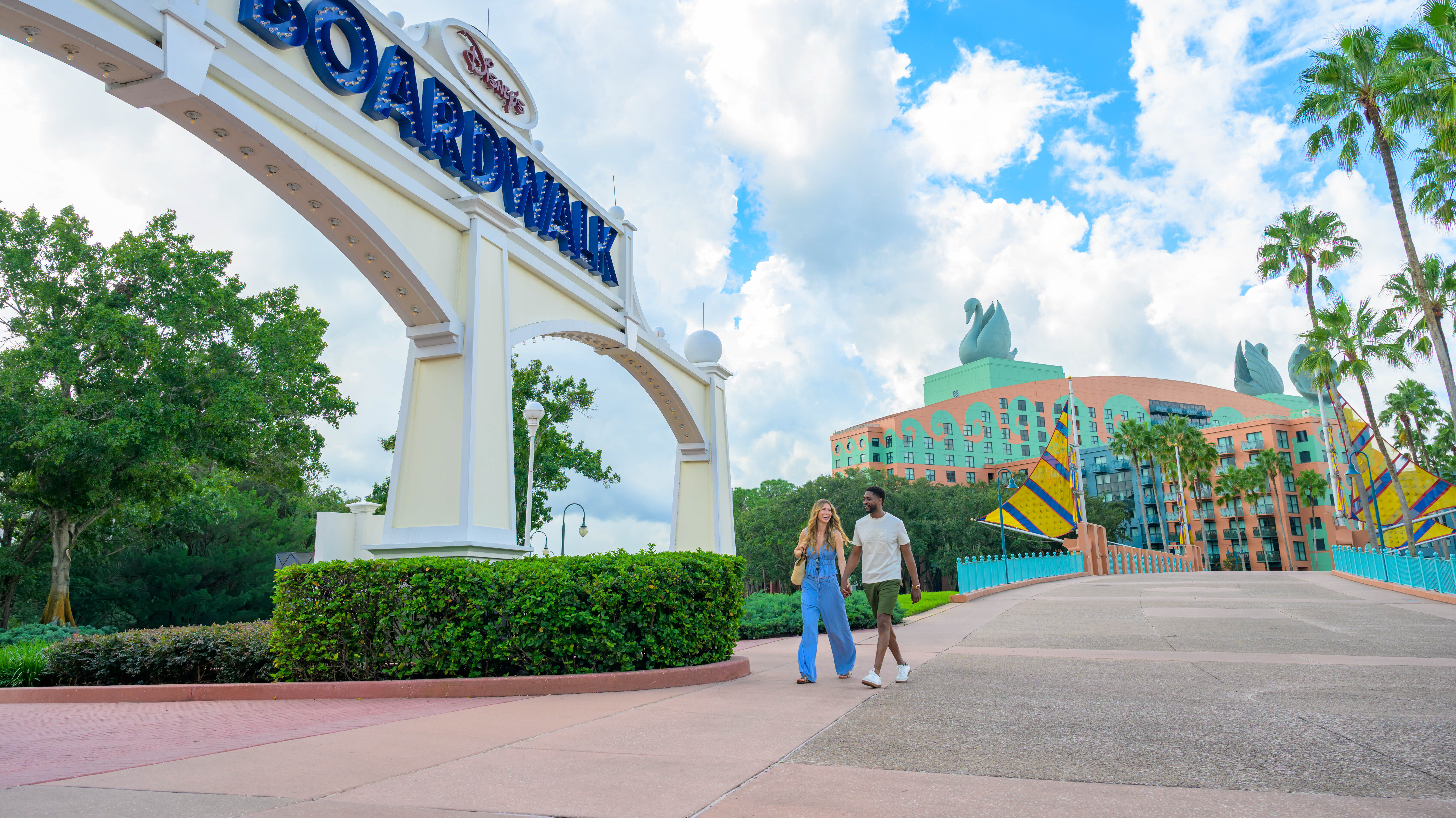 A couple walks together on a path beneath the Disneys BoardWalk entrance sign, with the swan-topped resort building in the background.