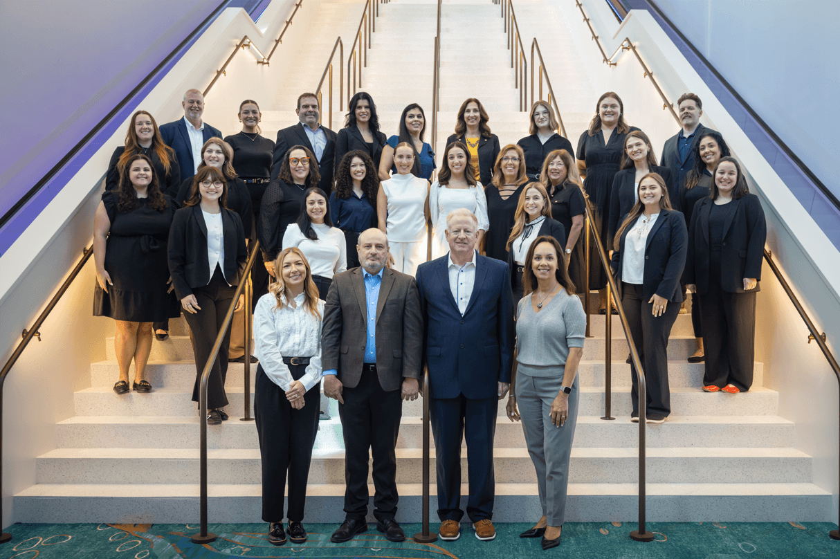 A group of 27 people from the Events Group dressed in business attire, pose for a photo on a staircase. Most are standing on the steps, while a few are in front at the base. The setting appears to be indoors, with bright lighting.