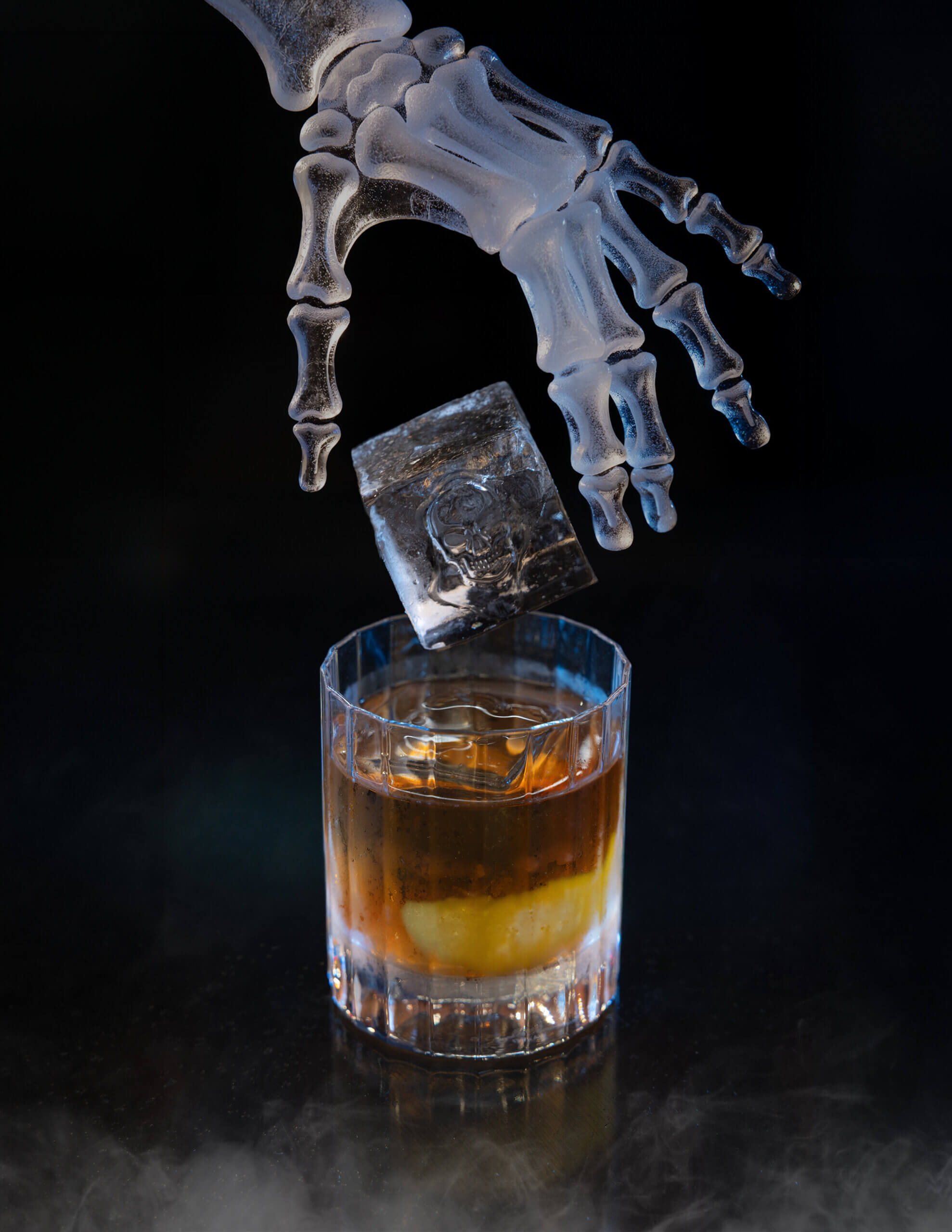Une main squelettique laisse tomber un glaçon dans un verre de whisky, sur un fond sombre et enfumé.
