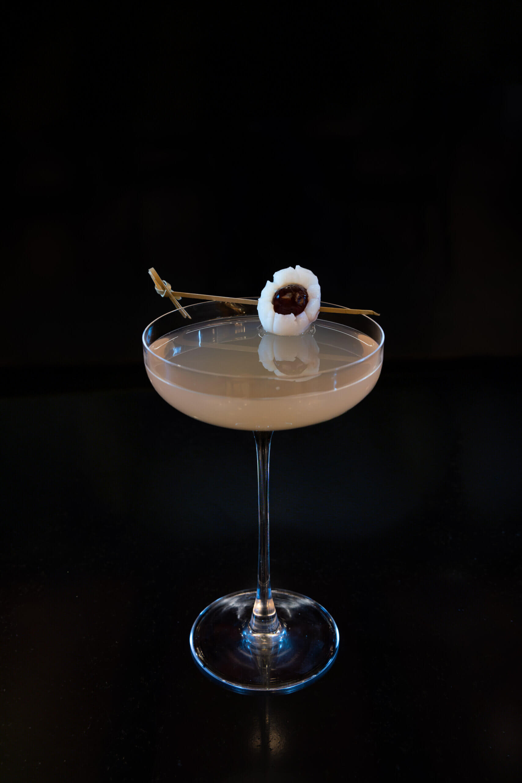 Un cocktail de couleur claire servi dans une coupe, agrémenté d'une brochette de litchi et de cerise, sur fond noir.