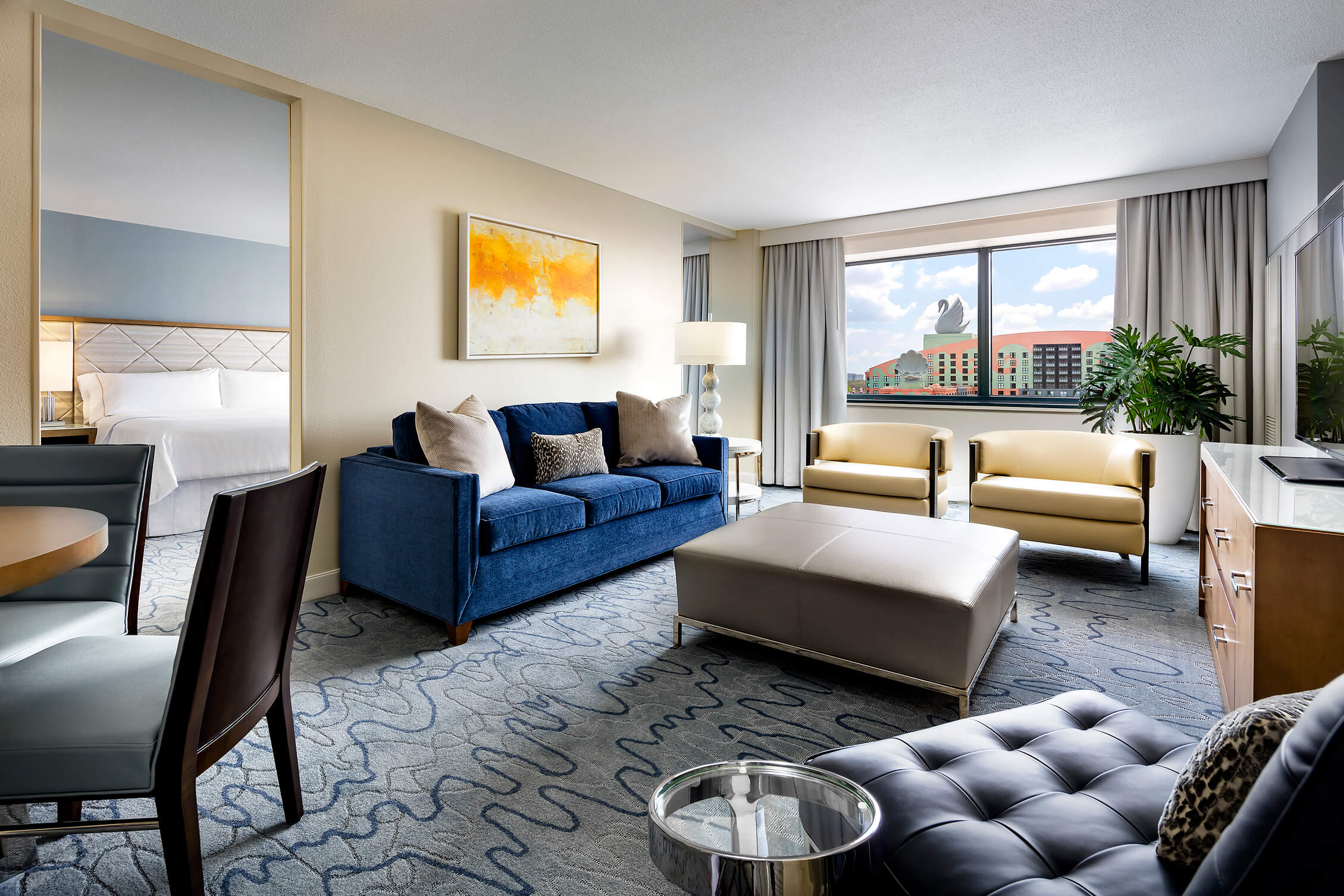 Suite de hotel con un sofá azul, dos sillones beige, una otomana de cuero y vistas a un complejo turístico en el que se divisa, a través de la ventana, una gran escultura de un cisne.