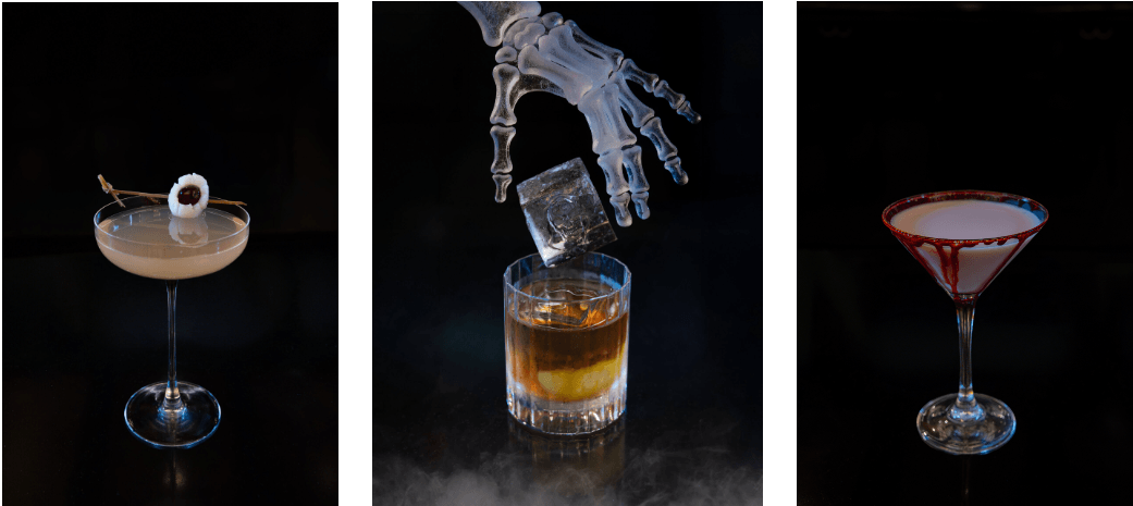 Three Halloween-themed cocktails: a drink garnished with an edible eye, a whiskey glass with an ice cube being dropped by a skeleton hand, and a martini glass with a red-stained rim.