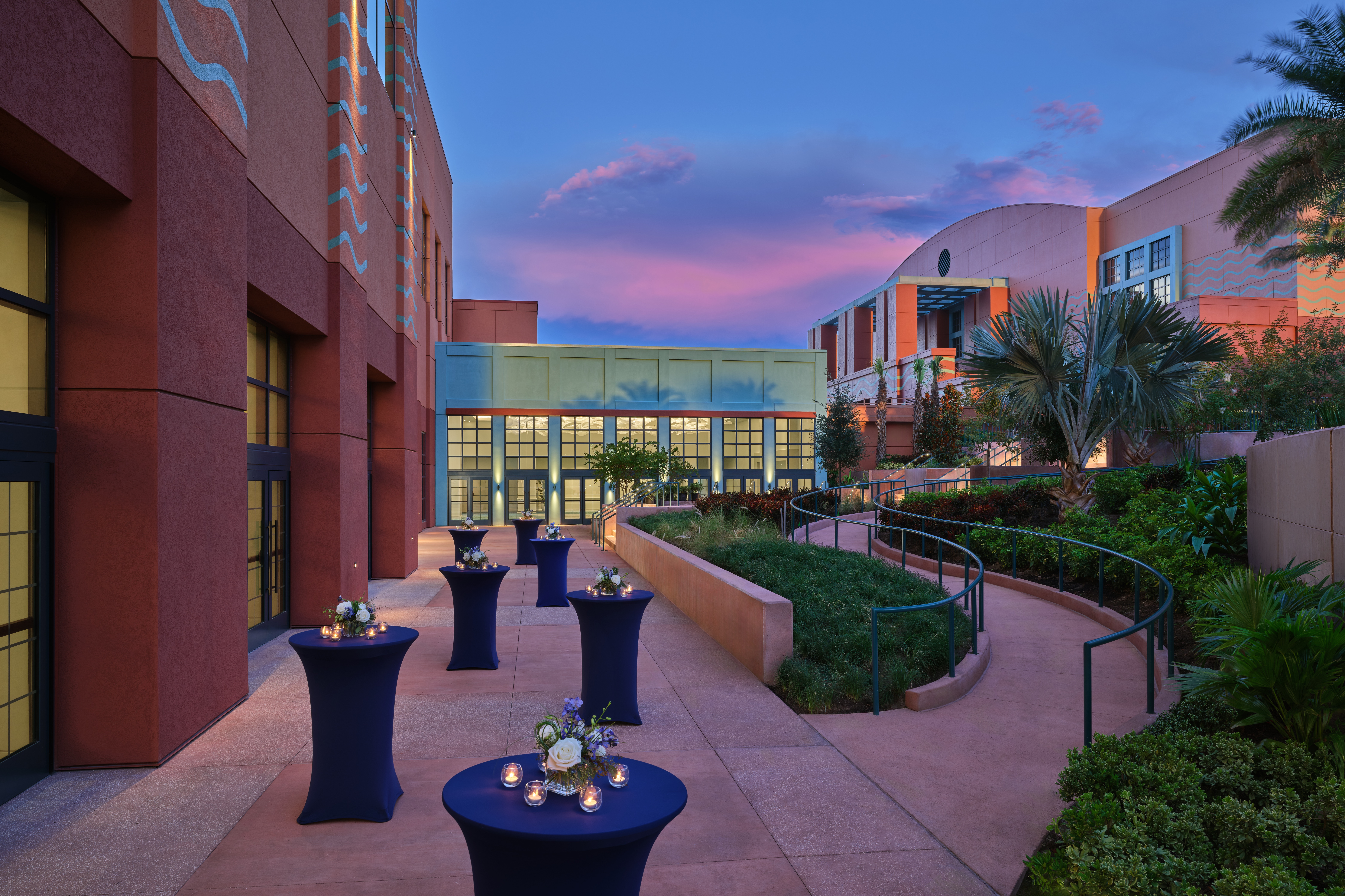 Outdoor patio at dusk with high-top tables covered in navy cloth and floral centerpieces, surrounded by modern buildings, greenery, and a purple-blue sky.
