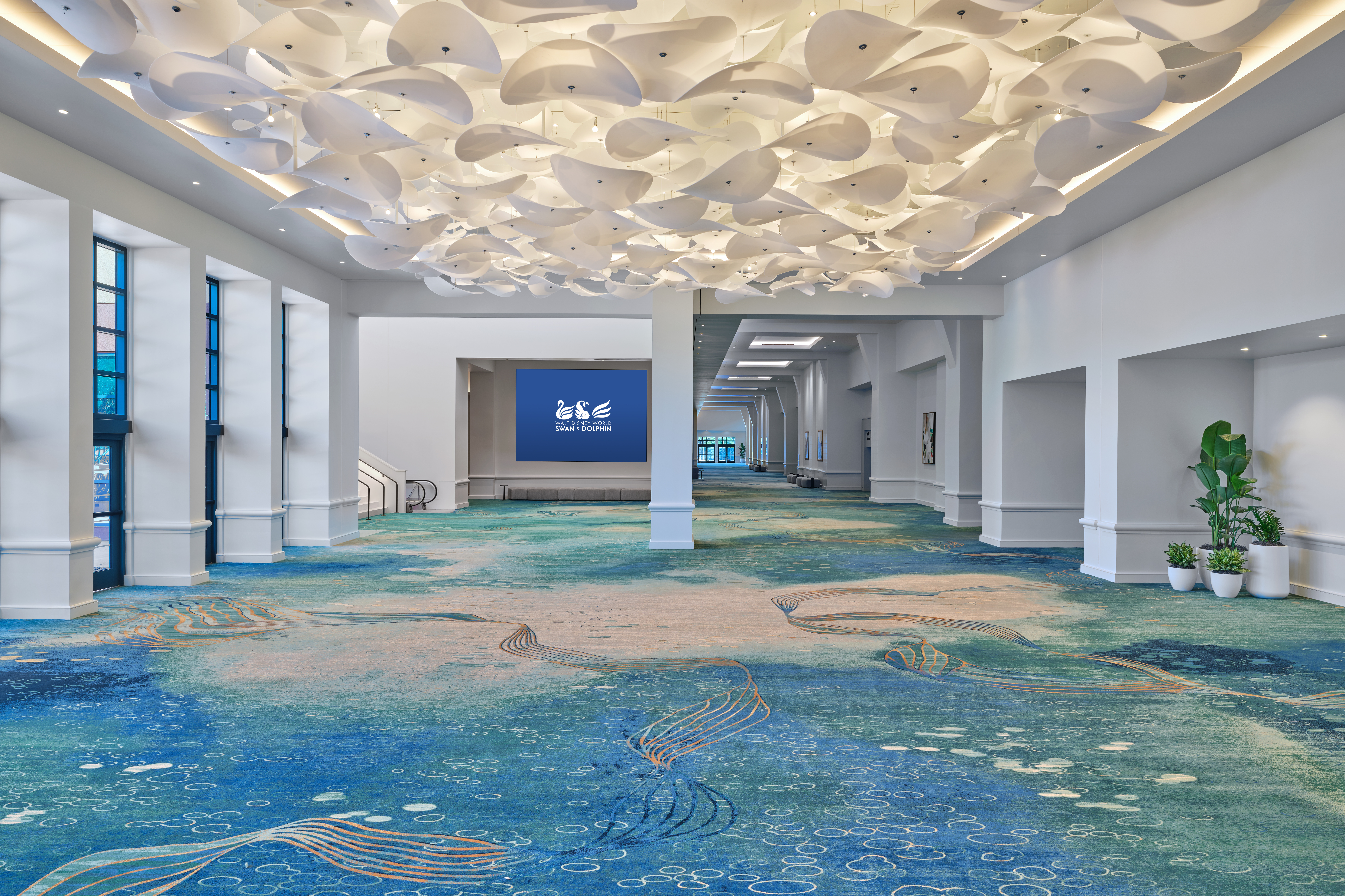Spacious, modern hallway with a blue-green patterned carpet, white sculptural ceiling fixtures, large windows on the left, and potted plants on the right. A sign at the end of the hall reads ISLE.
