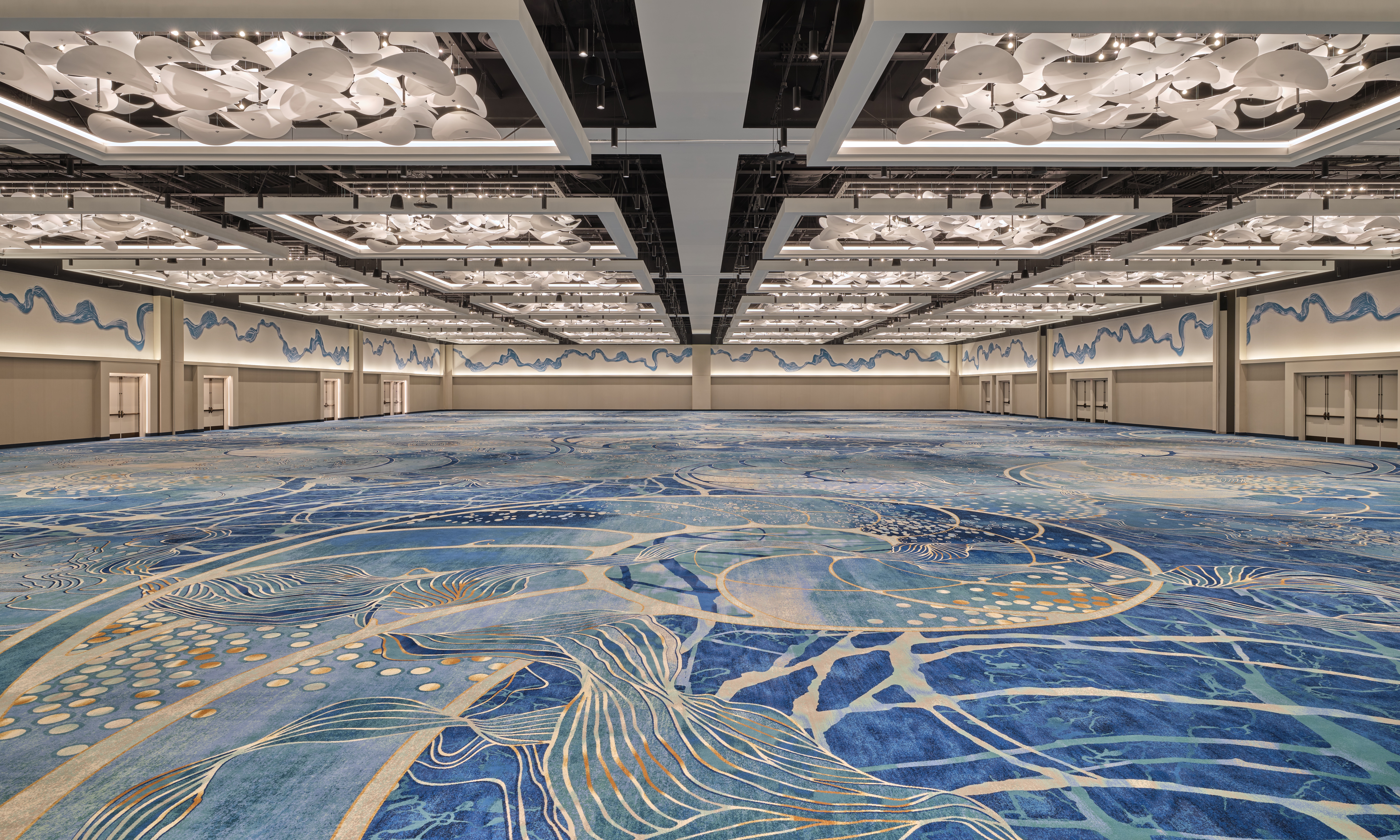 A large, modern ballroom with artistic blue carpeting, abstract ceiling lights, and wavy blue wall designs. The space is empty and expansive, with doors lining the far wall.