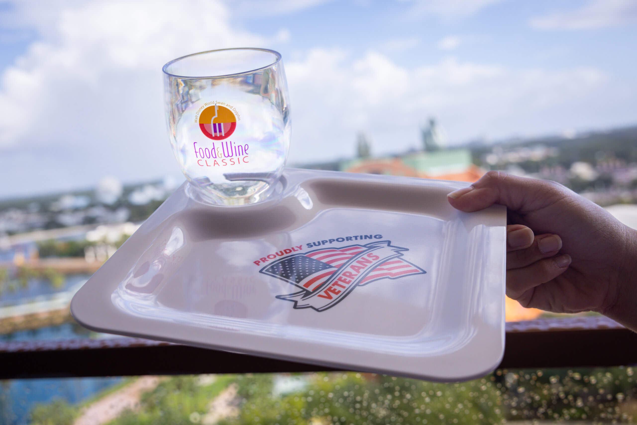Uma mão segura uma bandeja com uma bandeira americana e a frase “Proudly Supporting Veterans” (Orgulhosamente Apoiando os Veteranos), além de um copo com a inscrição “ Food &amp; Wine Classic ”, tendo como pano de fundo um cenário ao ar livre desfocado.