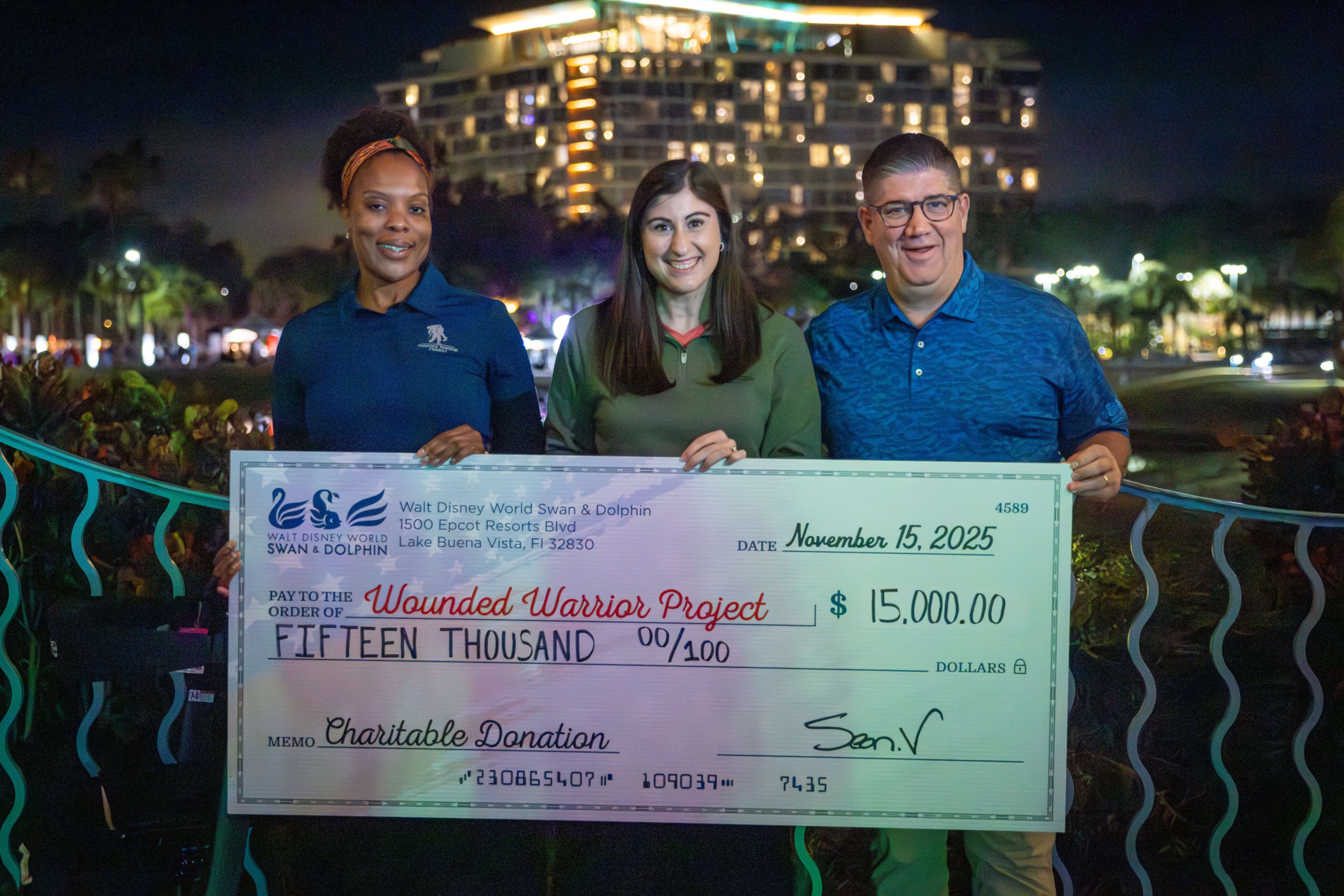 Três pessoas estão do lado de fora à noite, sorrindo e segurando um cheque gigante no valor de US$ 15.000, emitido em nome do Wounded Warrior Project como doação beneficente. Ao fundo, vê-se um hotel iluminado.