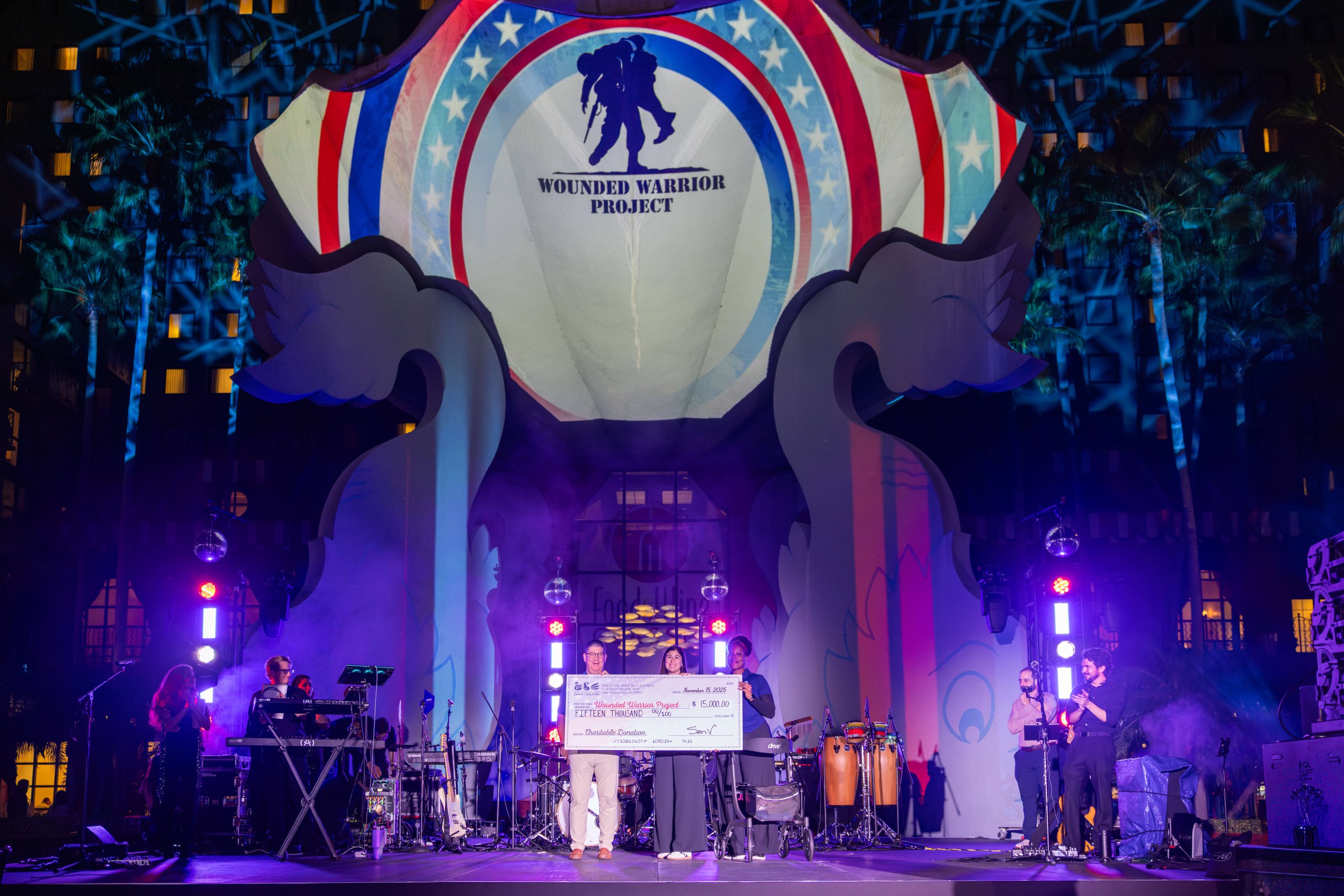 Um grupo está no palco segurando um cheque gigante diante de um grande cenário com o logotipo do Wounded Warrior Project, iluminado por luzes coloridas, com uma banda tocando ao vivo de cada lado.
