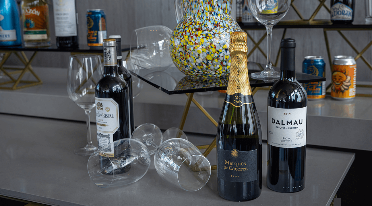 Tre bottiglie di vino e quattro bicchieri da vino vuoti sono disposti su un piano di lavoro grigio accanto a un vaso di vetro colorato e screziato, mentre sullo sfondo, sugli scaffali, sono esposti vari altri vasi e bicchieri.