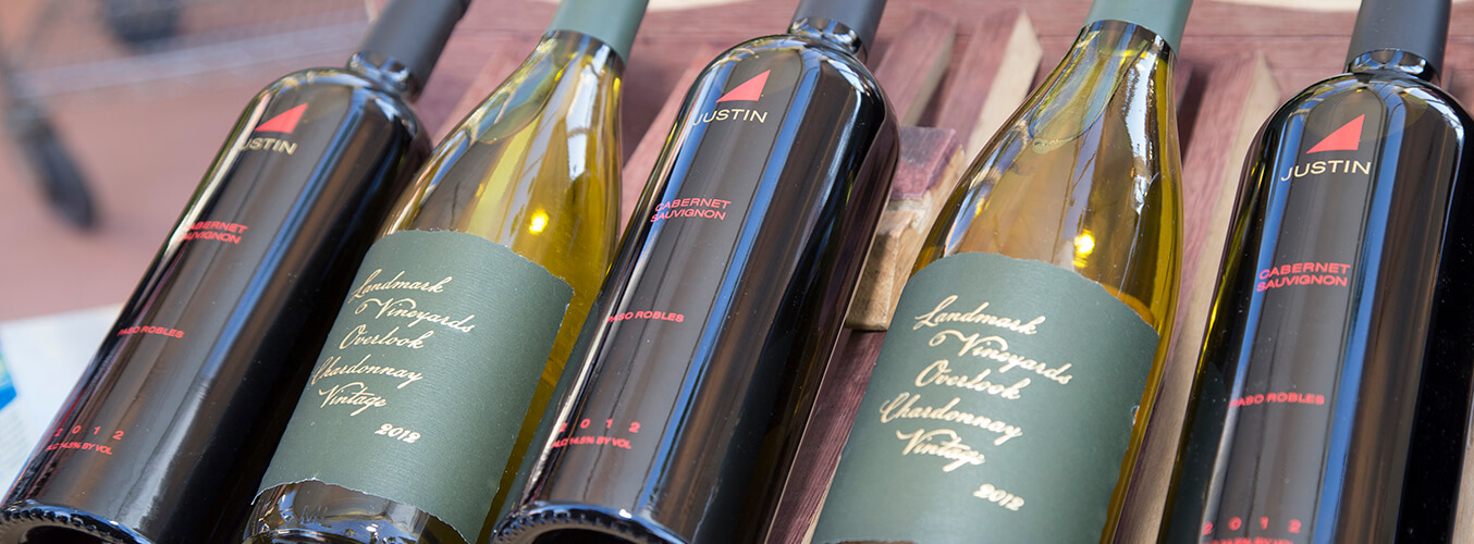 Пять бутылок вина, в том числе «Justin Cabernet Sauvignon» и «Landmark Vineyards Overlook Chardonnay», выставлены рядом на деревянной поверхности. Бутылки расположены под углом так, что видны этикетки.