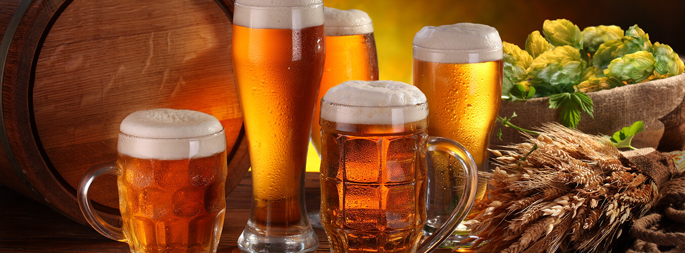 Five glasses of beer with frothy heads sit on a wooden table beside a barrel, a basket of hops, and a bundle of wheat, all set against a warm, glowing background.