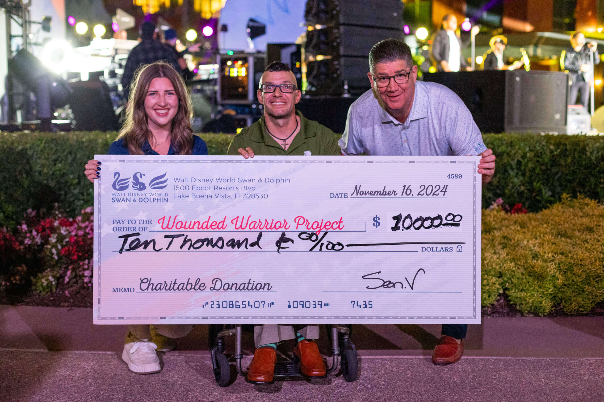 Três pessoas sorriem e seguram um grande cheque simbólico no valor de US$ 10.000, em nome do Wounded Warrior Project, datado de 16 de novembro de 2024, em um evento com luzes de palco e artistas ao fundo.