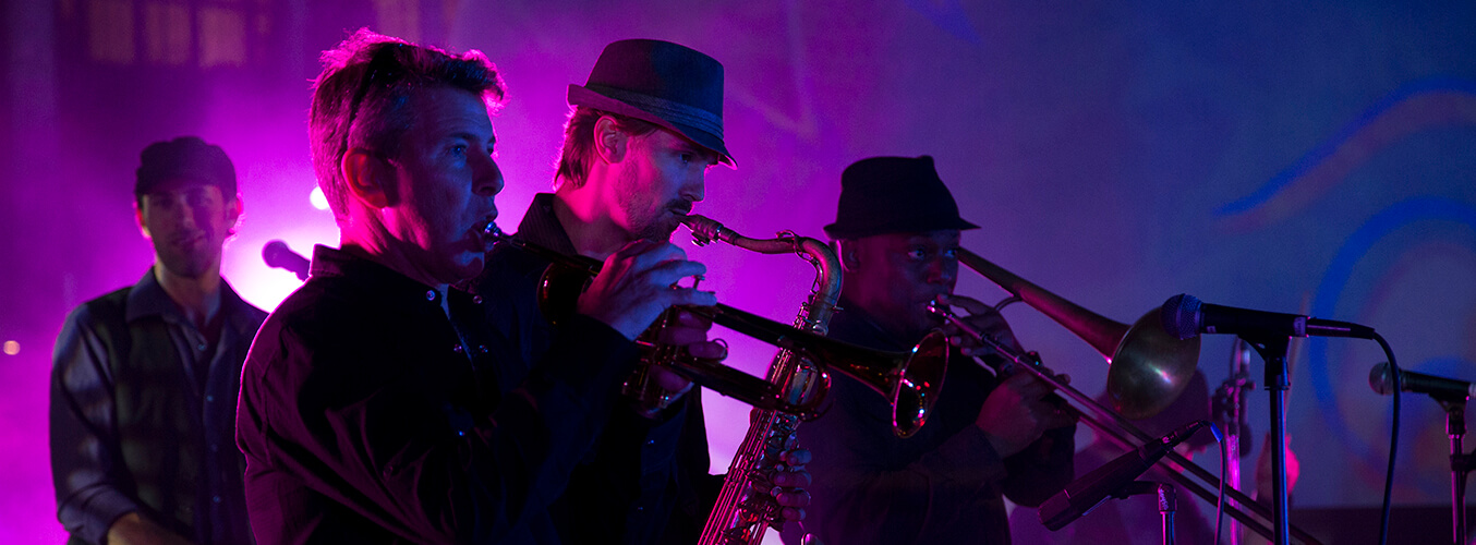 Four musicians perform on stage with brass instruments, including trumpet, saxophone, and trombone, under vibrant purple lighting. The background is dark and colorful, creating a lively concert atmosphere.