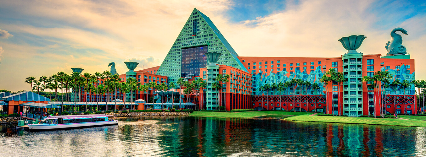 A colorful resort building with a large triangular central tower, giant decorative swans, palm trees, and a boat docked on a calm lake under a partly cloudy sky.