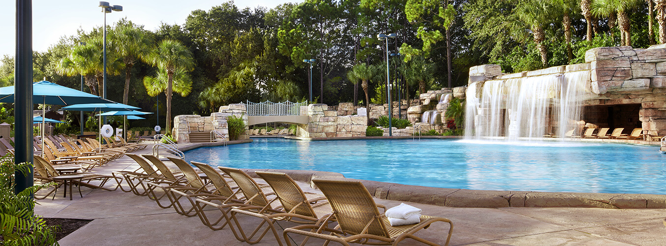 Outdoor swimming pool with rows of empty lounge chairs, blue umbrellas, a stone waterfall feature, a small bridge, and lush green trees in the background on a sunny day.