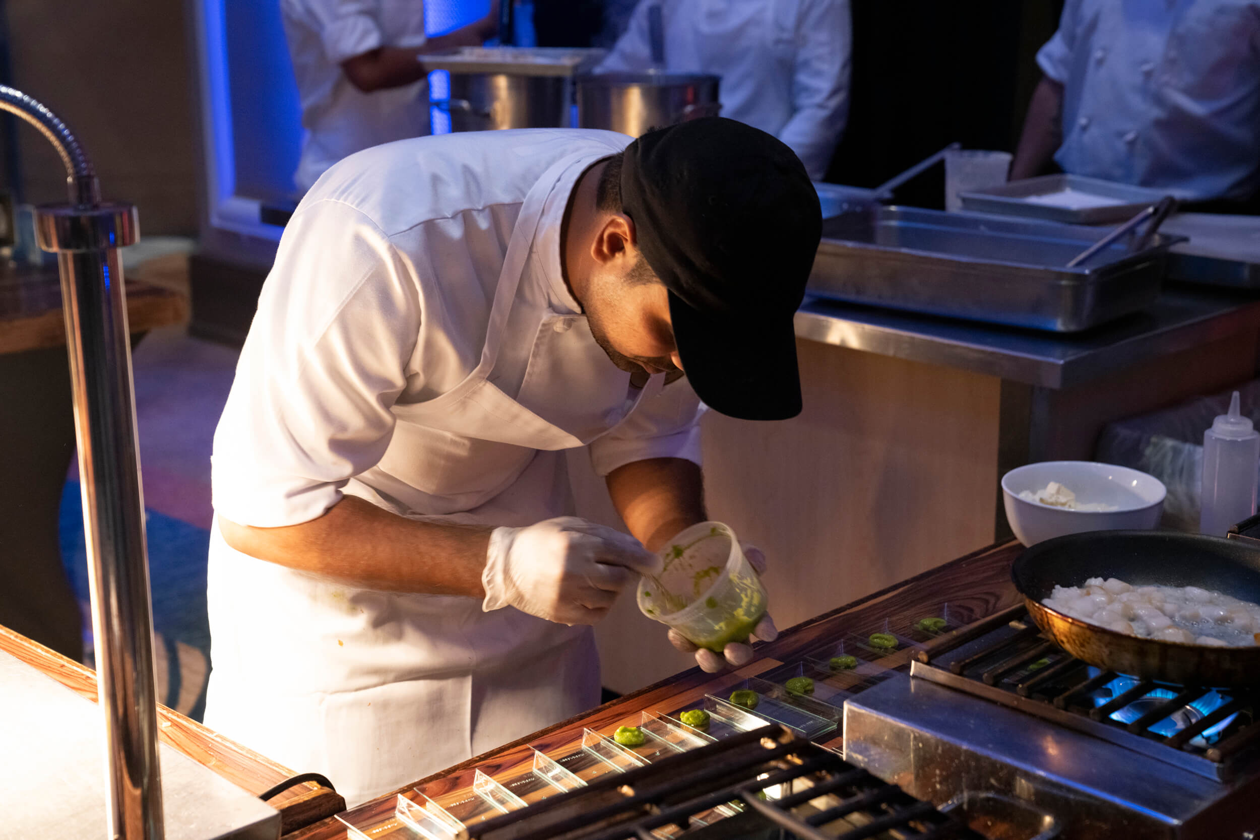 A chef wearing a white uniform and black cap carefully adds green sauce to small dishes on a counter, working in a well-lit kitchen with cooking pans and supplies around him.