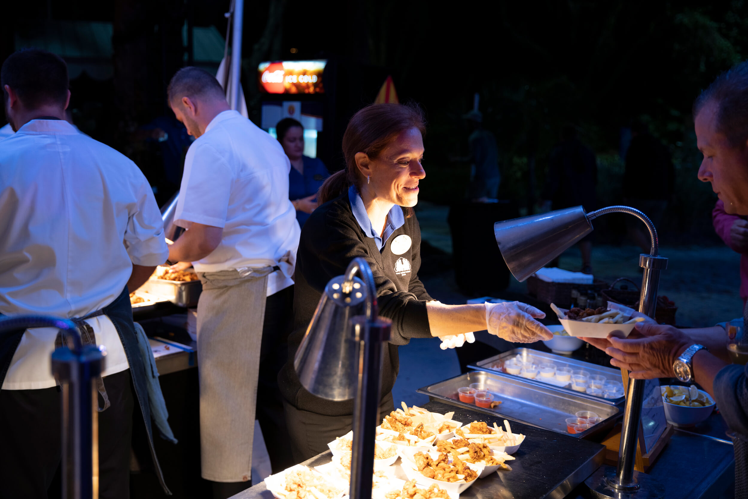A smiling server hands food to a guest at an outdoor night event, with other staff preparing dishes at a well-lit serving station filled with trays of food. People are gathered around, enjoying the festive atmosphere.