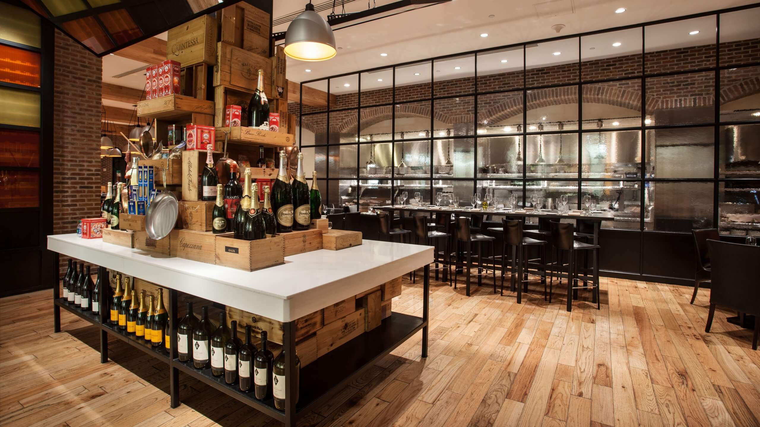 A modern restaurant interior featuring a central display of stacked wine and champagne bottles on a white table, wood flooring, brick walls, and a glass wall revealing an industrial-style kitchen.