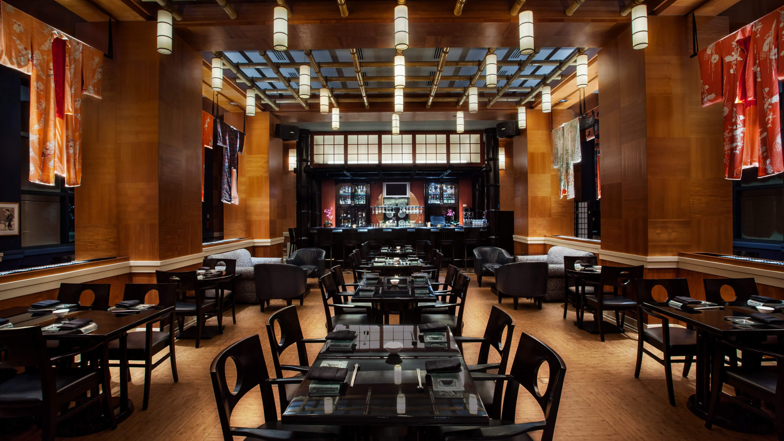 A modern restaurant with wooden walls, black tables and chairs, hanging lights, and kimonos displayed on the walls. A bar is visible at the back, and the space is warmly lit with a sophisticated atmosphere.