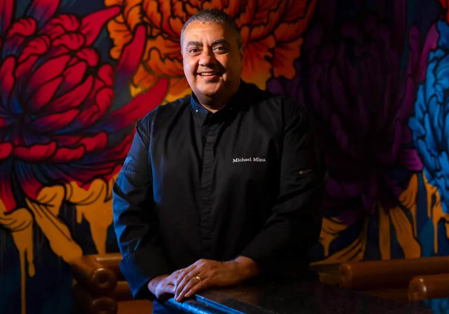 A smiling chef in a black coat with Michael Mina embroidered on it stands in front of a vibrant, colorful floral mural.