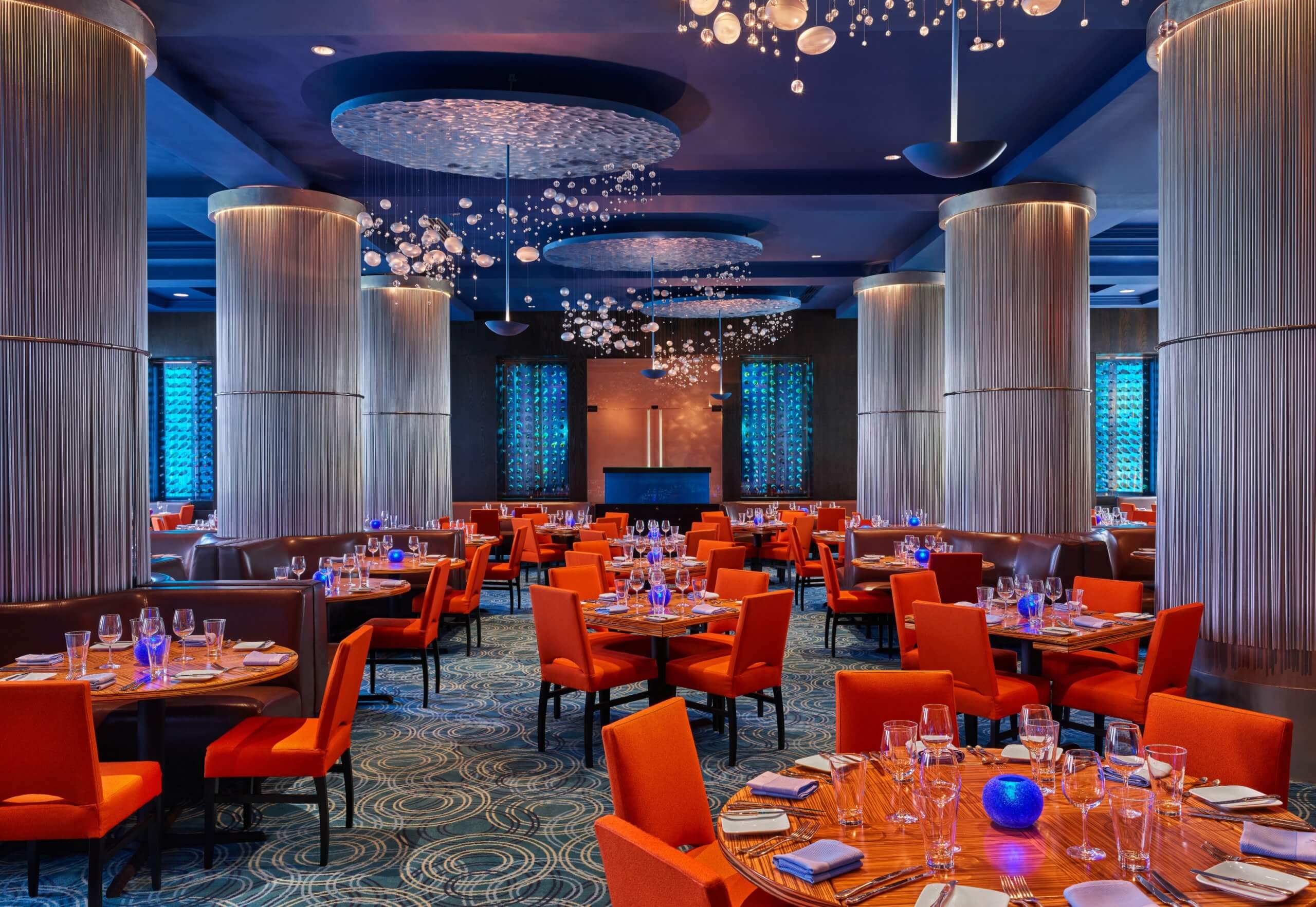 A stylish restaurant interior with round wooden tables, orange chairs, and elaborate ceiling lights. Tall columns and blue accent lighting create a modern, elegant atmosphere. Tables are set with glassware and napkins.