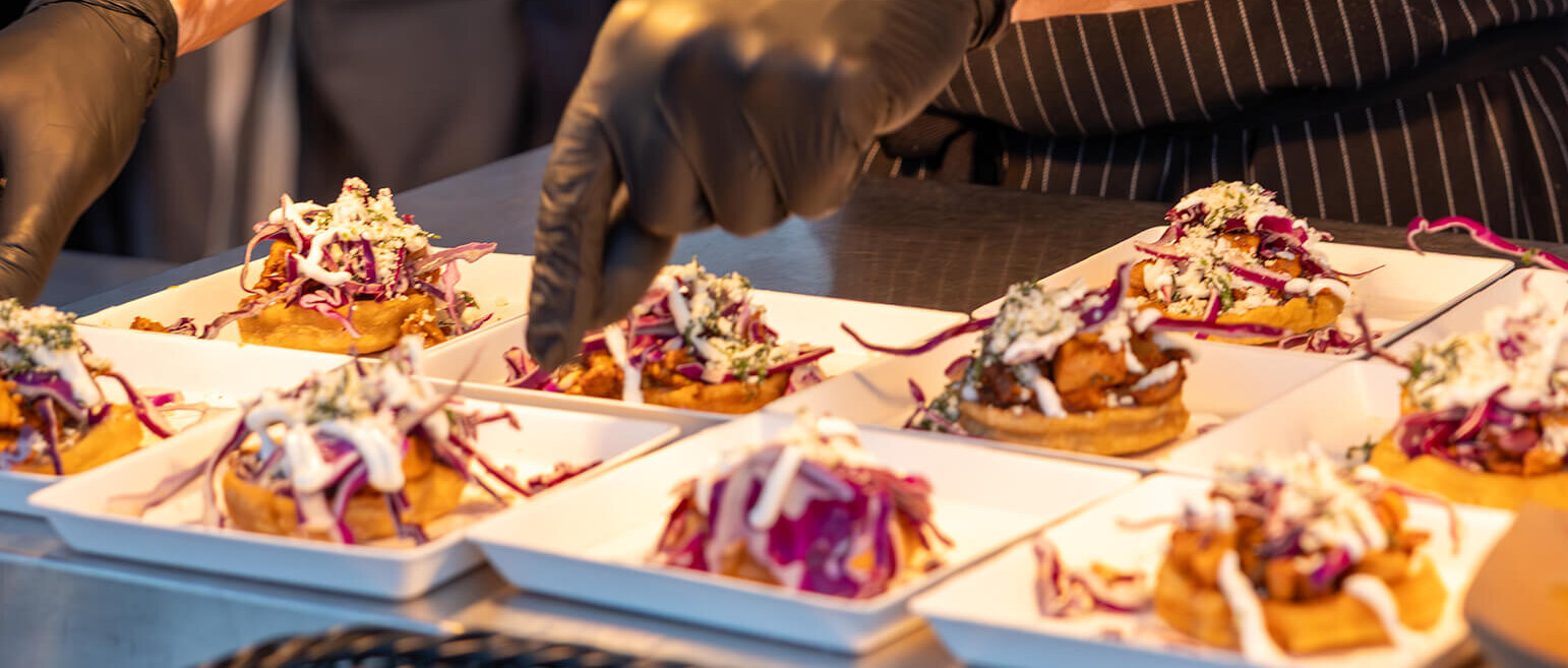 A chef wearing black gloves and a striped apron prepares and arranges plates of food topped with shredded red cabbage, sauce, and herbs on a counter.