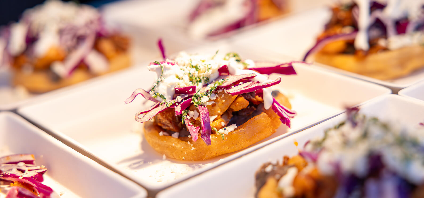 A close-up of a small, round savory dish topped with shredded red cabbage, crumbled cheese, herbs, and diced vegetables, served on a white plate with similar dishes in the background.