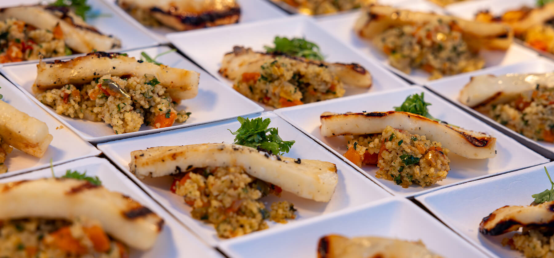 Rows of square white plates, each with grilled fish fillets and a serving of mixed vegetable couscous, topped with a fresh parsley garnish. The plates are neatly arranged for catering or a buffet.