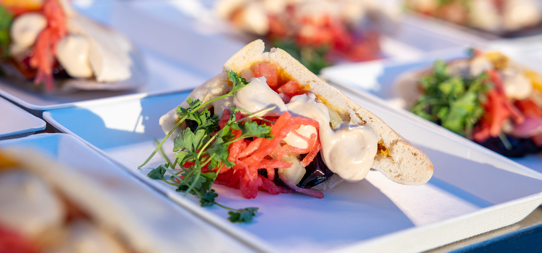 A pita sandwich filled with fresh vegetables, pink pickled onions, herbs, and creamy sauce is served on a white tray, with more similar pita sandwiches in the background.