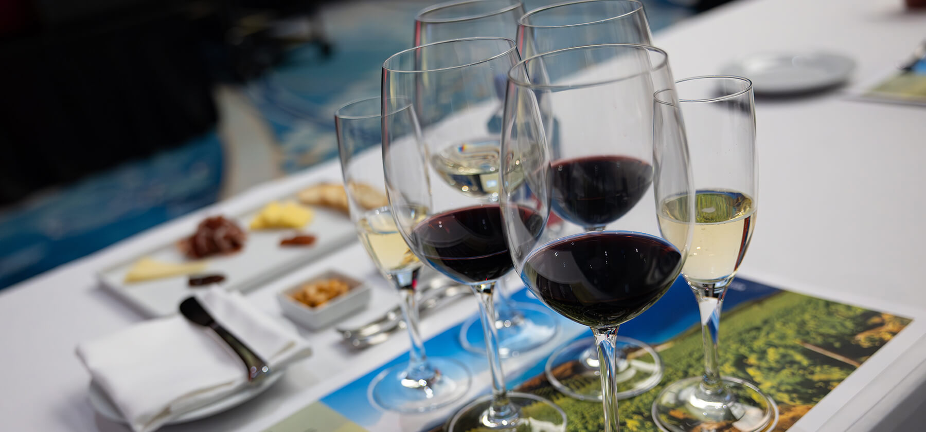 Cinque bicchieri da vino, tre pieni di vino rosso e due di vino bianco, sono disposti su un tavolo con, sullo sfondo, piatti di formaggio, frutta secca e cracker, a suggerire un evento di degustazione di vini.