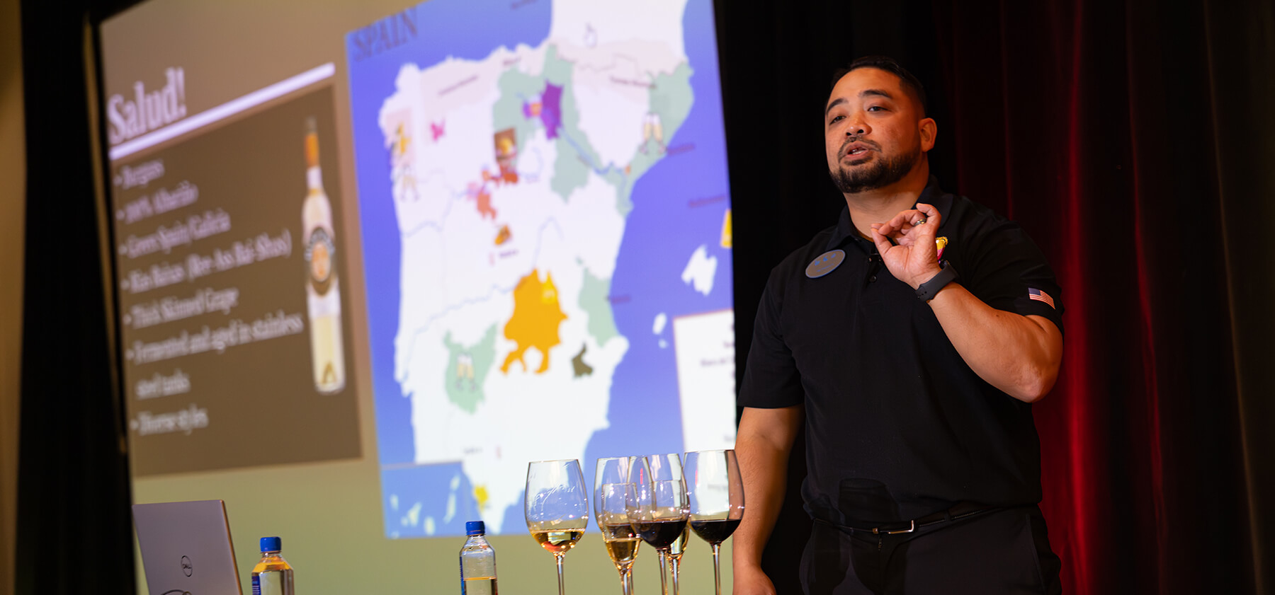 A man gives a presentation on Spanish wine, gesturing as he stands beside a screen showing a map of Spain and wine regions. Several wine glasses and water bottles are on a table in front of him.
