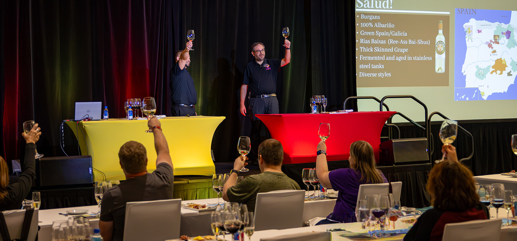 A group of people seated at tables raise wine glasses toward two presenters on stage, who are also holding up glasses. A presentation slide about Spanish wine is displayed in the background.