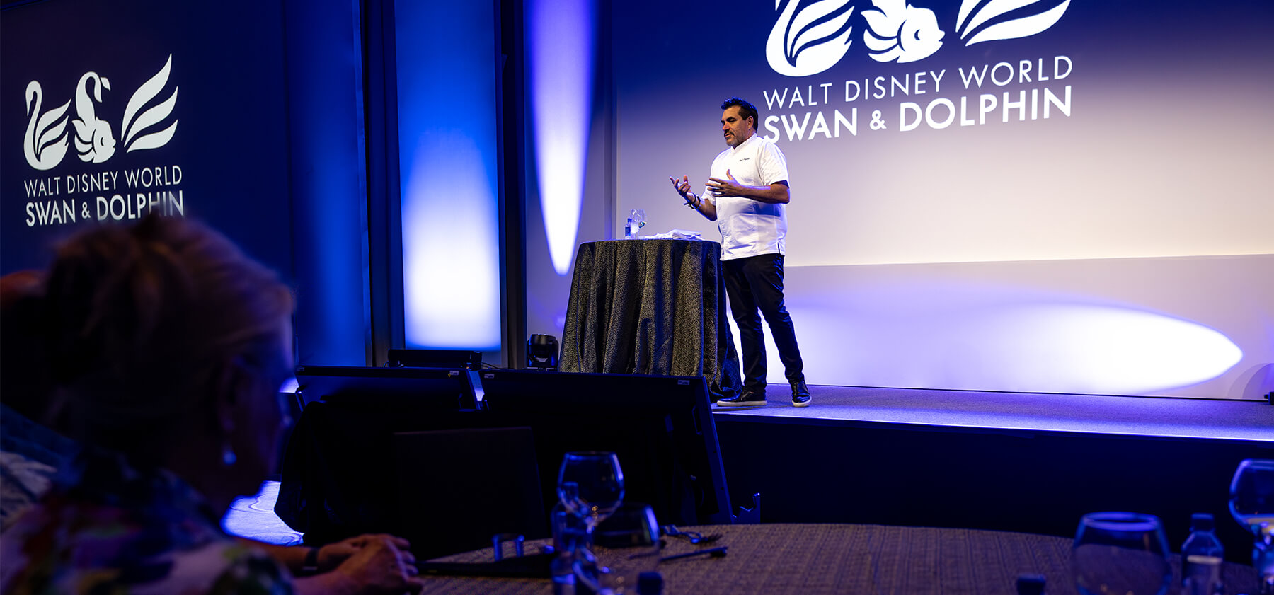 Todd English stands and speaks on stage at a Walt Disney World Swan & Dolphin event, with a logo projected behind him. Audience members are seated at tables in the foreground.