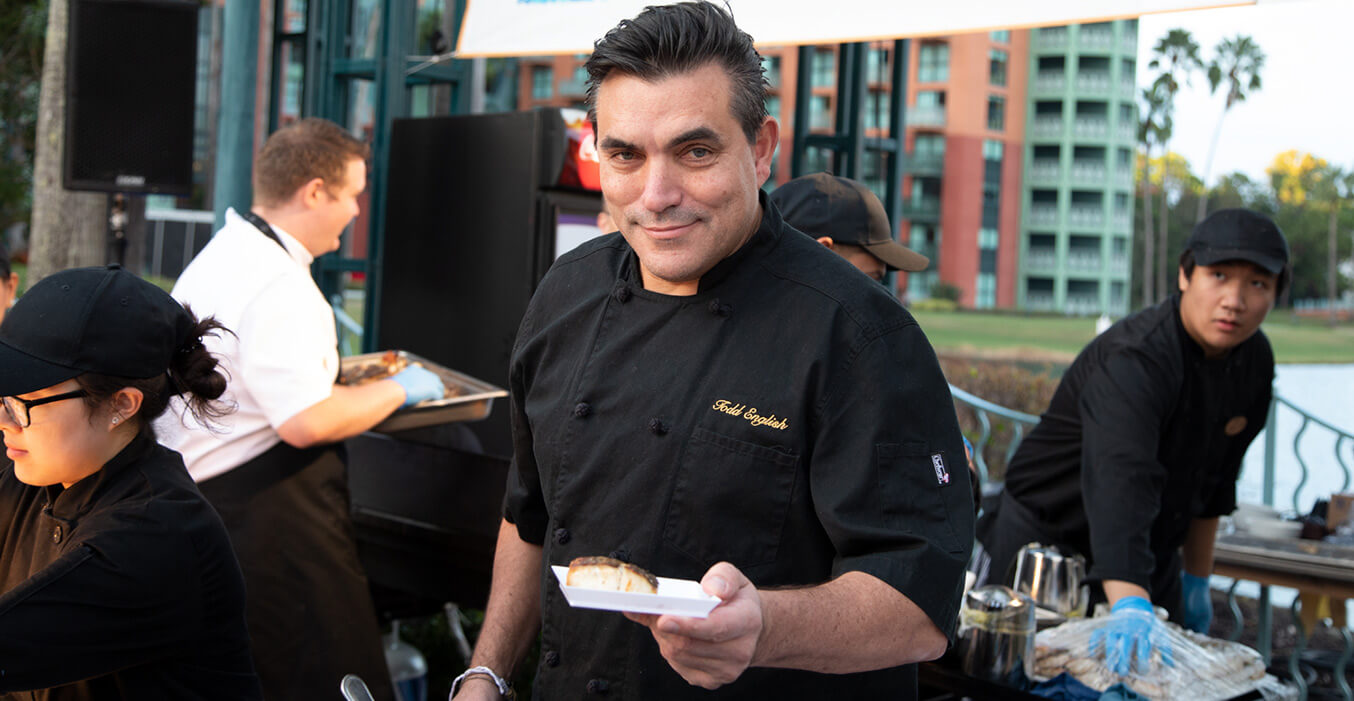 A chef in a black uniform holds a plated dish and smiles at the camera, while other staff members prepare food at an outdoor event near a waterfront and tall buildings.