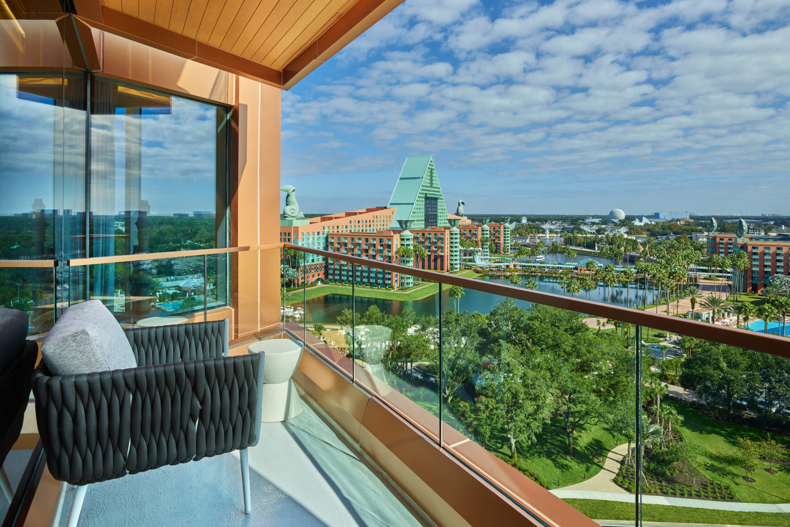 A hotel balcony with two black woven chairs and a small table overlooks a scenic view of modern buildings, a pool, green trees, and a partly cloudy blue sky.