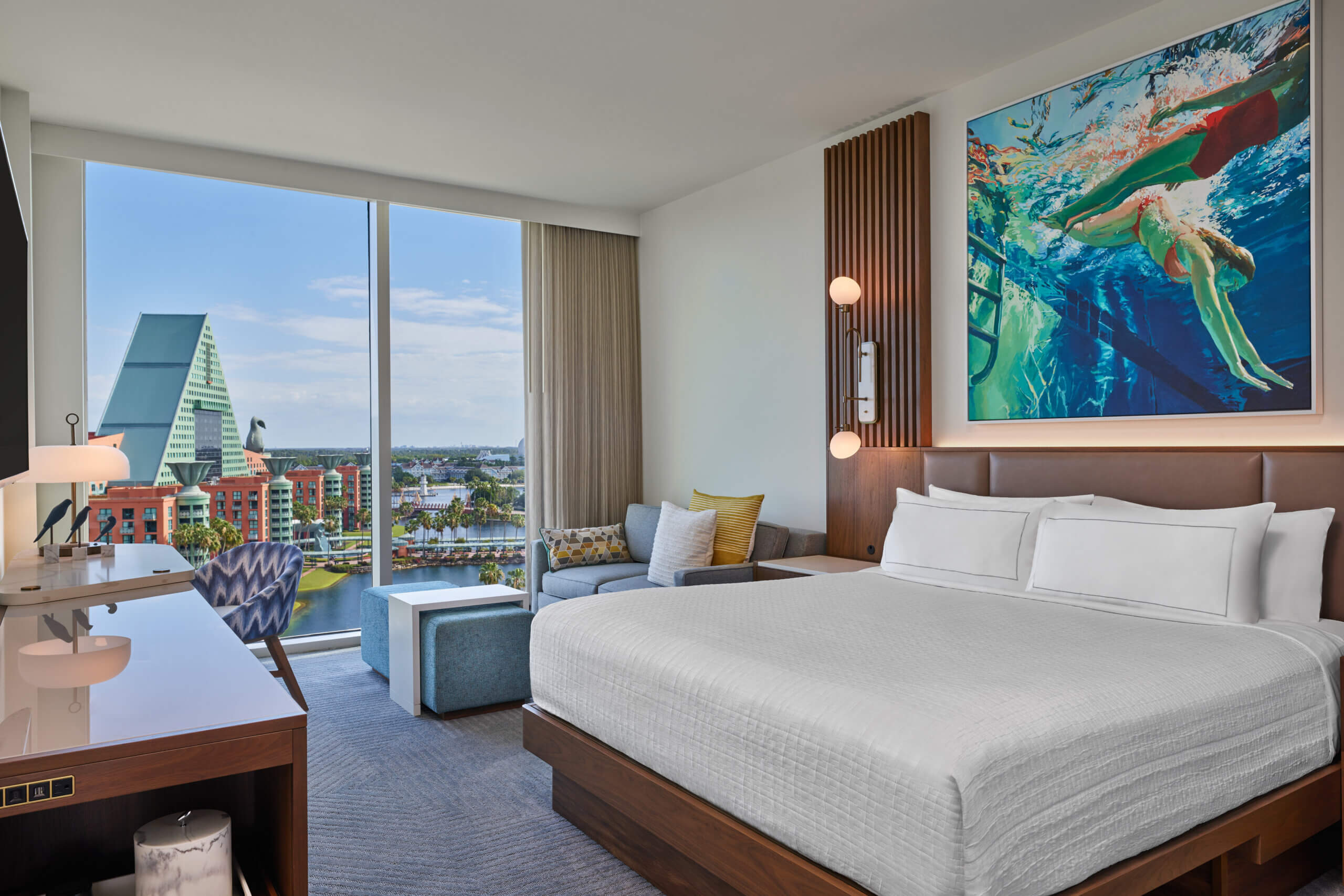 A modern hotel room with a large bed, desk, couch, and vibrant underwater-themed artwork. Floor-to-ceiling windows offer a view of a unique triangular building and a cityscape outside.