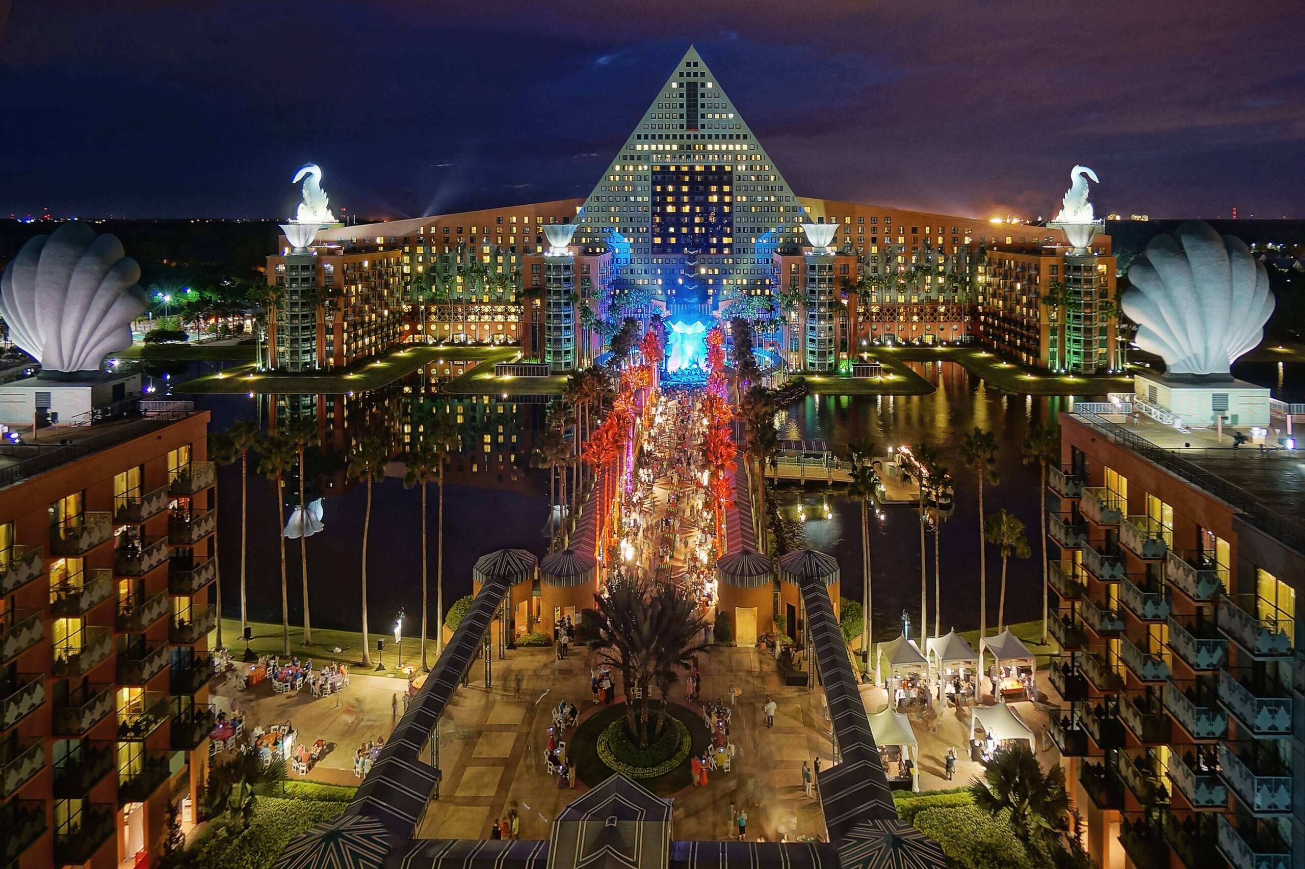 Nighttime view of the illuminated Walt Disney World Dolphin Hotel, featuring a pyramid-shaped center, two large fish sculptures, and palm tree-lined walkways with crowds and reflections in the water.