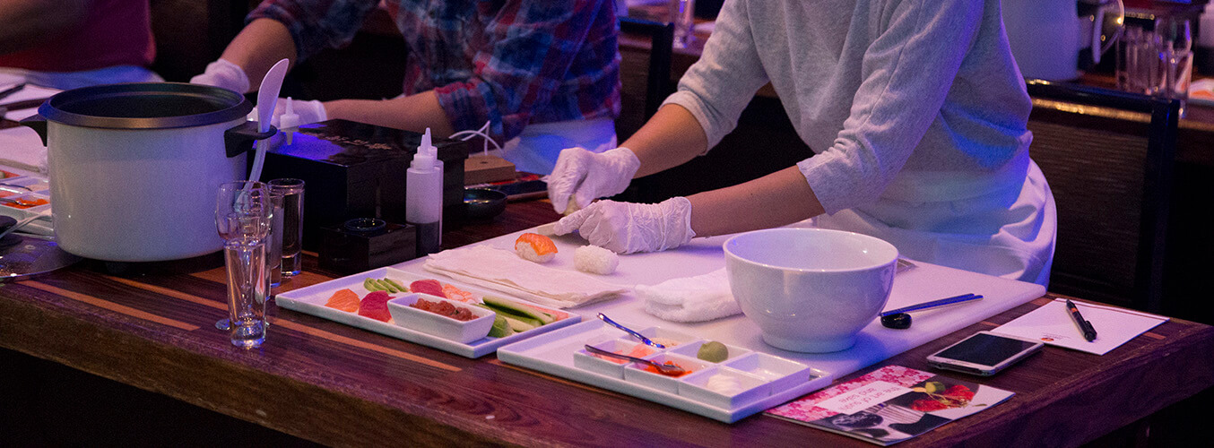 Two people wearing gloves prepare sushi at a counter with plates of fresh ingredients, a rice cooker, and kitchen tools under soft lighting. A bowl, chopping board, and recipe card are also on the table.