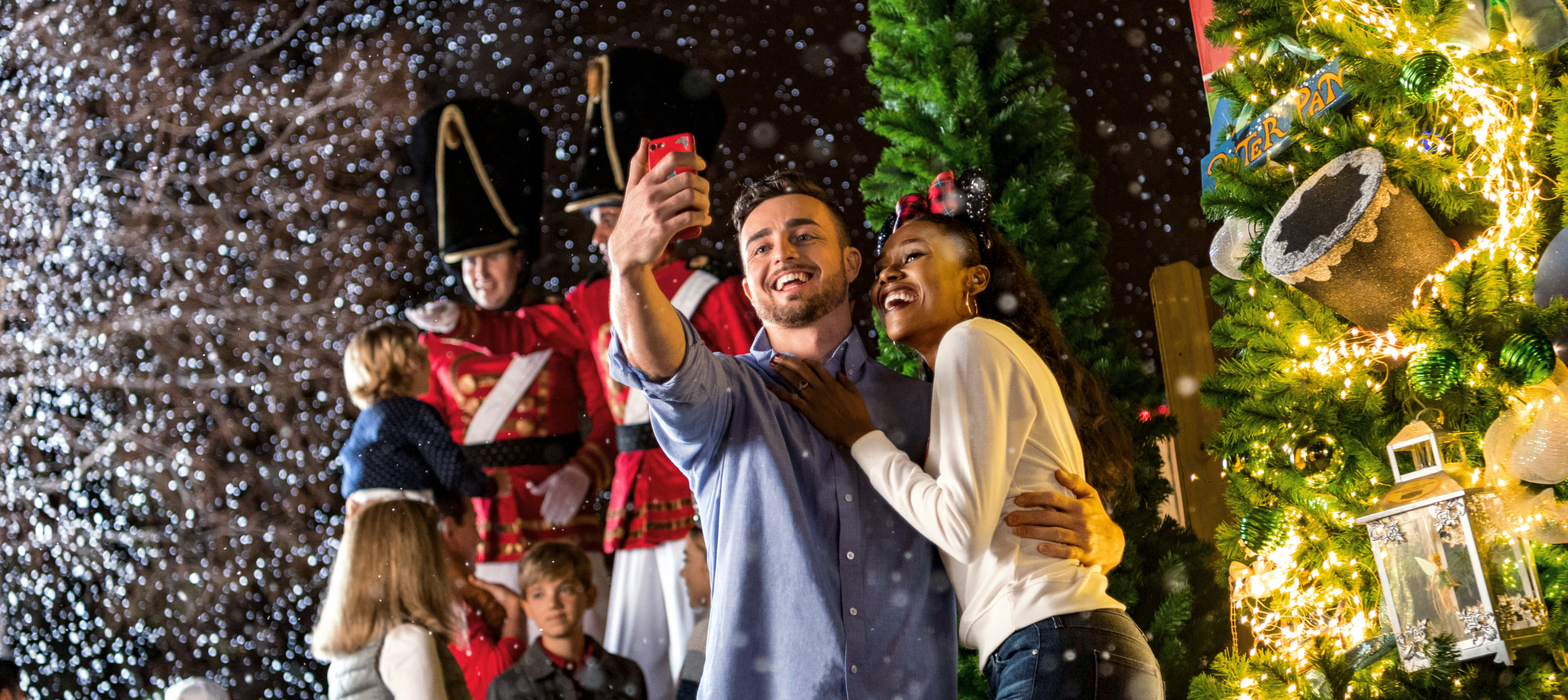 A couple smiles and takes a selfie in front of a decorated Christmas tree as artificial snow falls. People dressed as toy soldiers and children are visible in the festive, joyful scene.