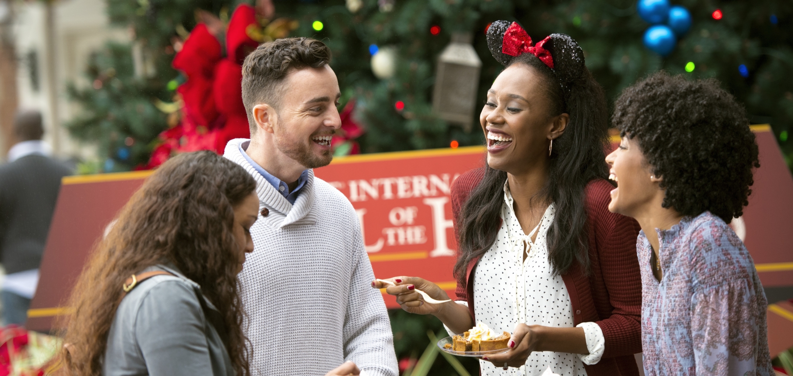 Four friends laugh and enjoy food together outdoors in front of a decorated Christmas tree. One woman wears festive Minnie Mouse ears, and they all appear happy and cheerful.