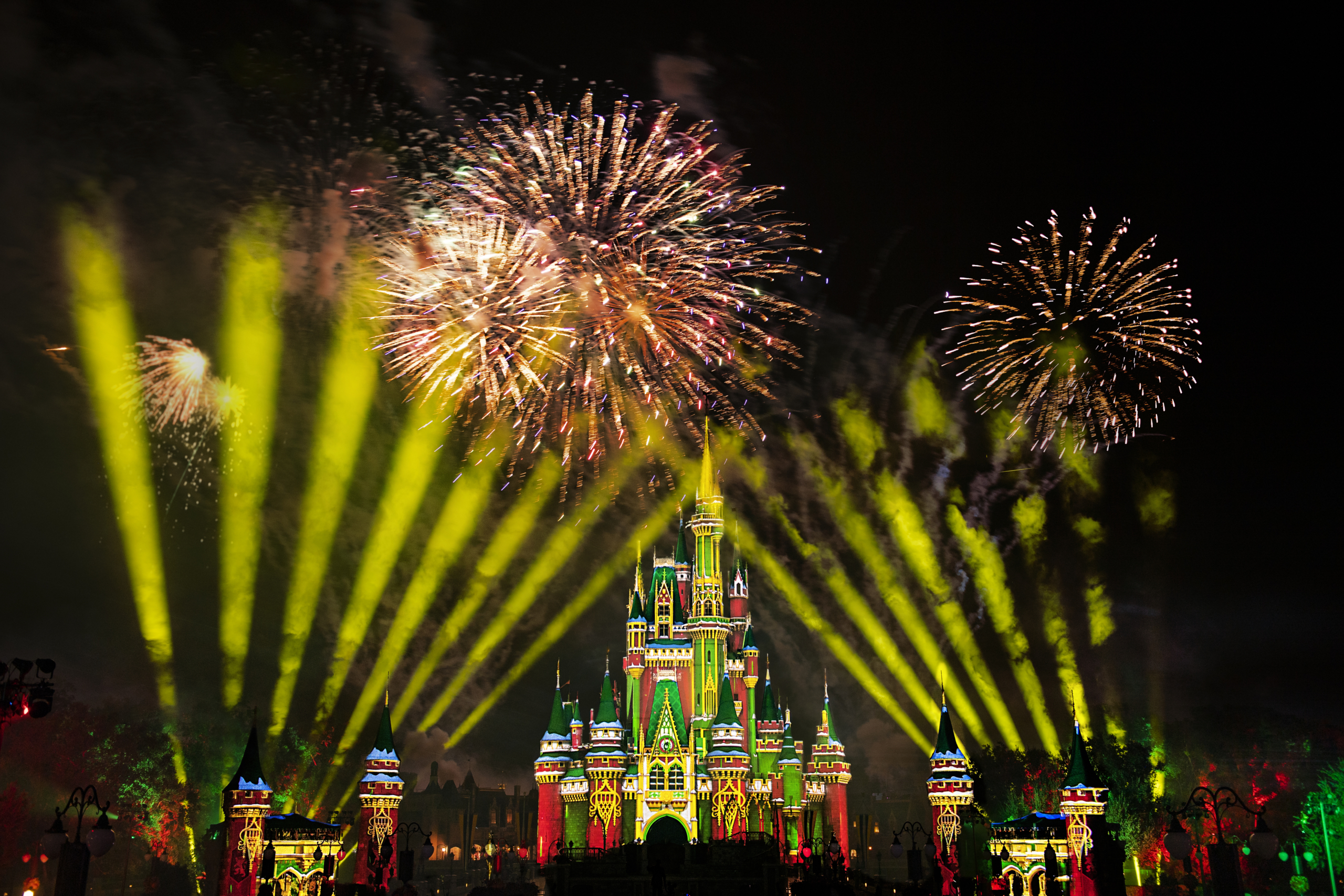 Colorful fireworks and yellow spotlights illuminate a brightly lit Disney castle at night, creating a festive and magical atmosphere.