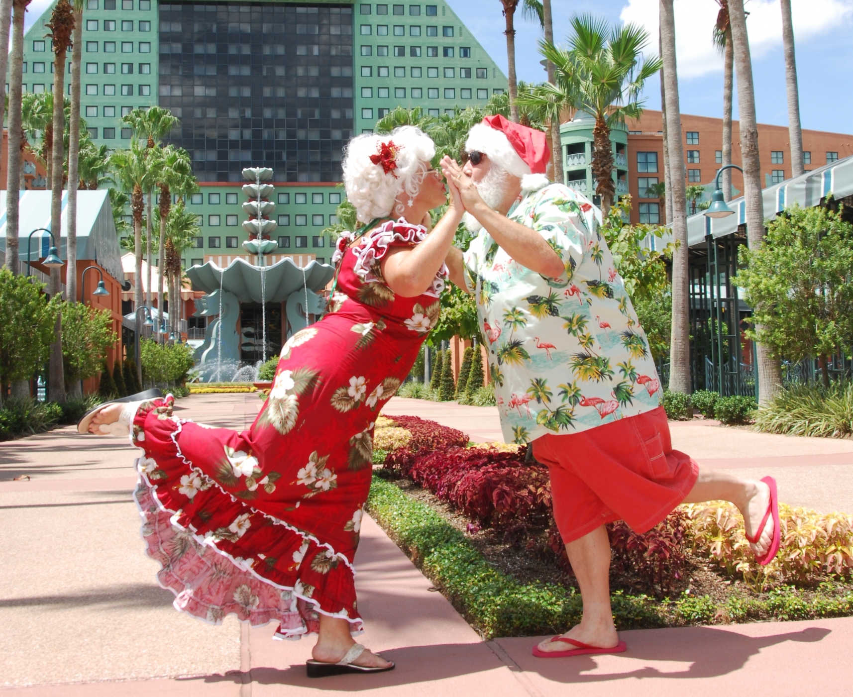Un couple de personnes âgées déguisées en Mère Noël et en Père Noël, vêtus de tenues de vacances aux couleurs tropicales, se tapent joyeusement dans la main tout en se tenant sur une jambe, dans un décor en plein air ensoleillé et bordé de palmiers.