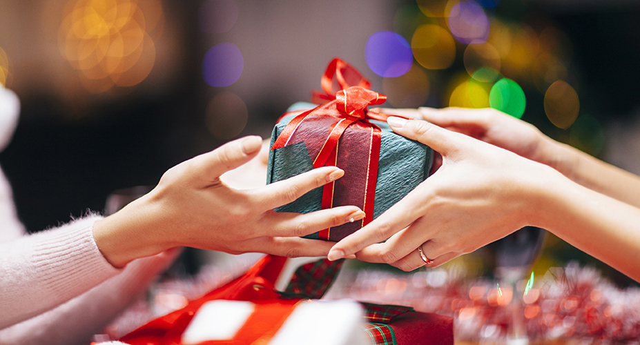 Two people exchanging a wrapped gift with a red ribbon, surrounded by festive decorations and blurred colorful lights in the background.