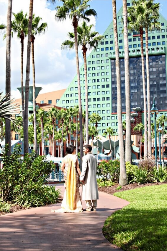 A couple dressed in traditional Indian attire walks hand in hand along a sunny, palm tree-lined path in front of a modern, pyramid-shaped hotel building.