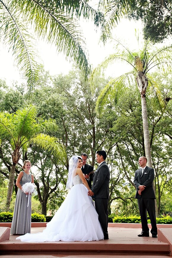 A bride and groom stand facing each other, holding hands, during an outdoor wedding ceremony with two attendants and an officiant under tall palm trees.