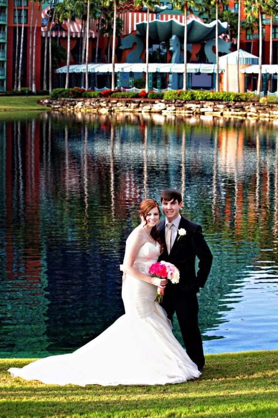 A bride in a white gown and groom in a black suit smile and pose together by a lakeside, holding a bouquet of pink and white flowers, with colorful buildings and palm trees reflected in the water behind them.