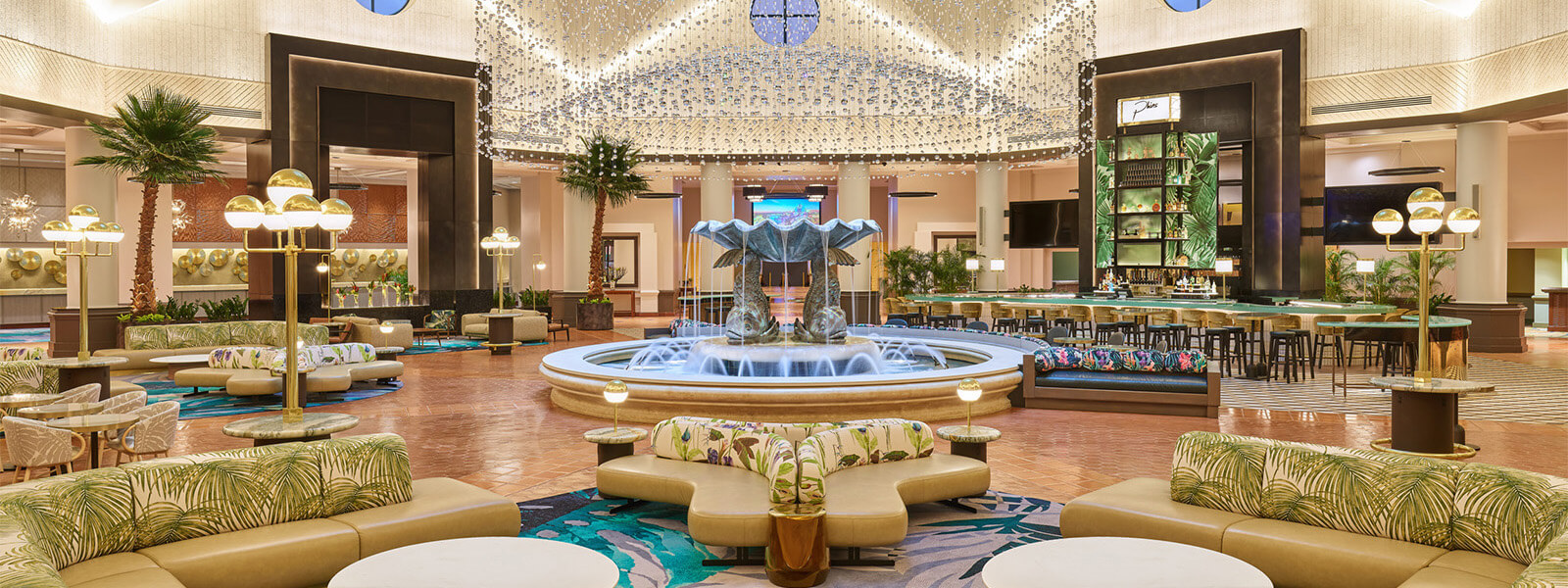 Spacious hotel lobby with a large central fountain, modern sofas, round tables, lush green plants, stylish lamps, and a well-lit bar area under a dome ceiling decorated with hanging lights.