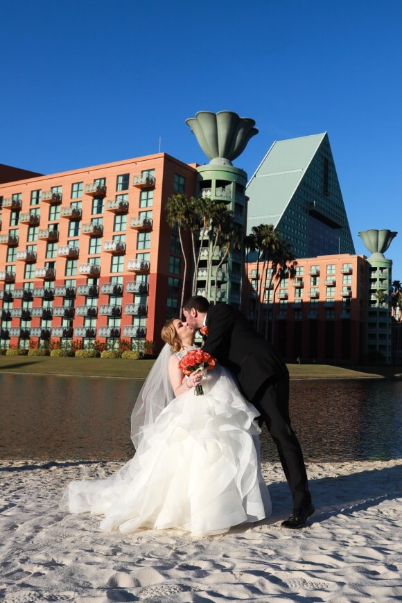 A bride and groom share a kiss on a sandy beach in front of a modern hotel with geometric shapes and palm trees, under a clear blue sky. The bride holds a bouquet of flowers and wears a white gown.